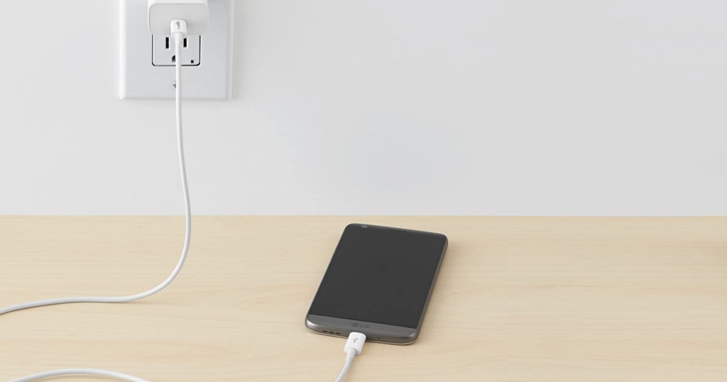 cell phone on wood table charging with white cord plugged into wall