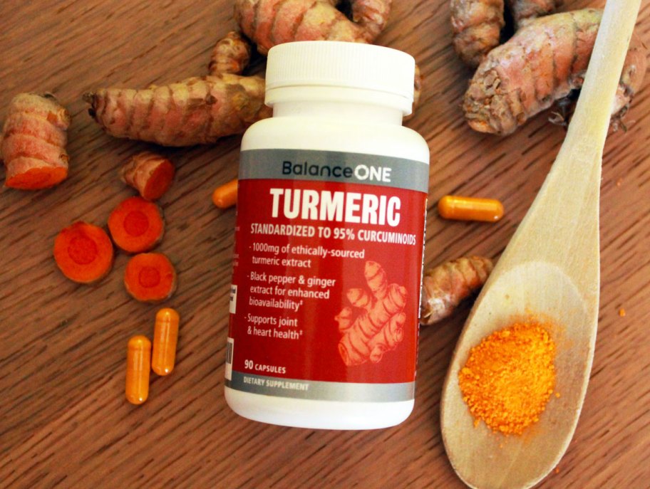 bottle of balance one tumeric capsules next to ginger root and spoon with turmeric on it
