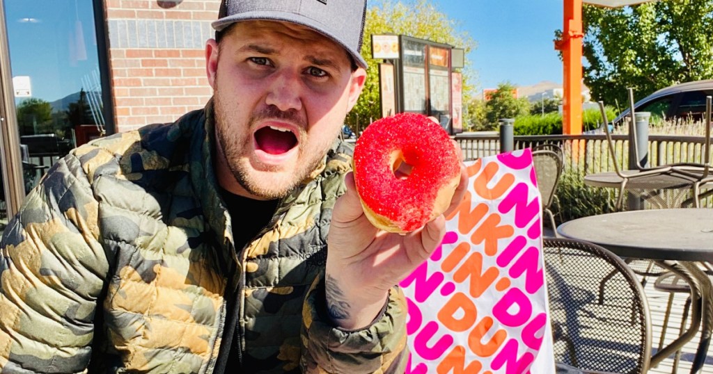 man sitting at table outside holding donut with red sprinkles