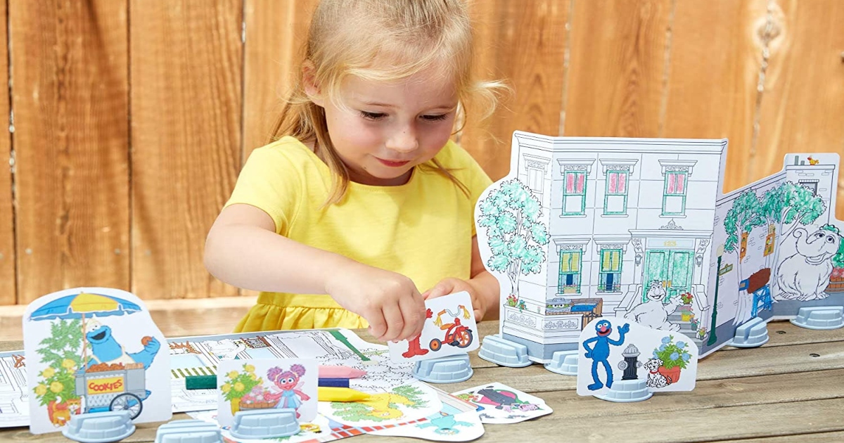 Girl in yellow shirt playing outside on wooden table with a Green Toys Coloring Set. The colored figures are propped up with plastic holders.