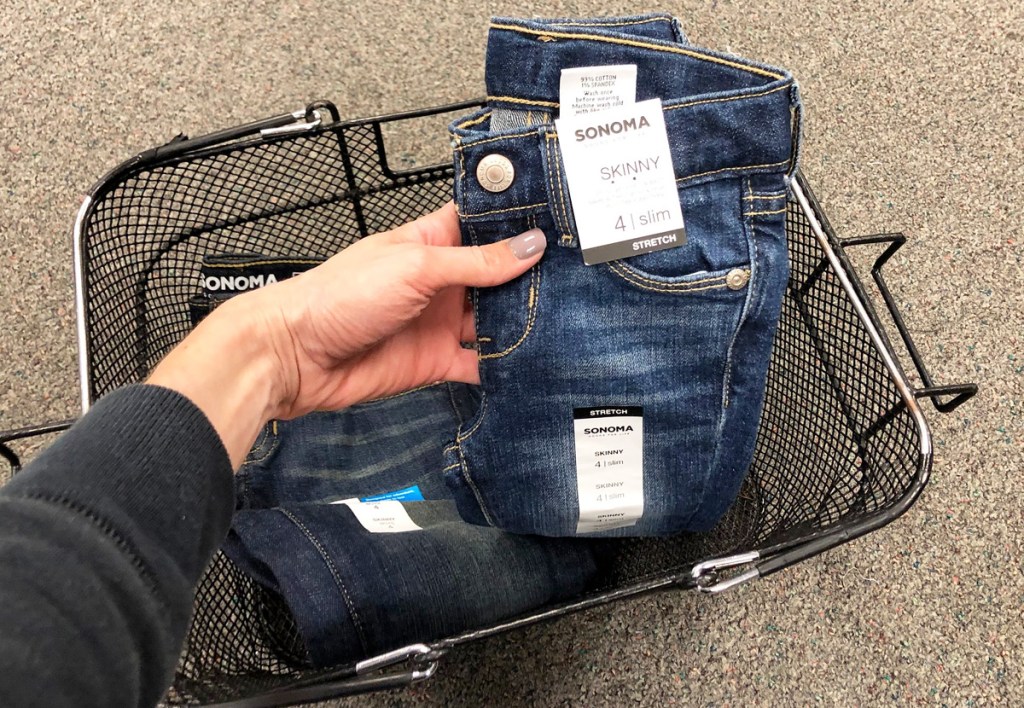 woman holding up a pair of sonoma brand girls jeans from black shopping basket on floor