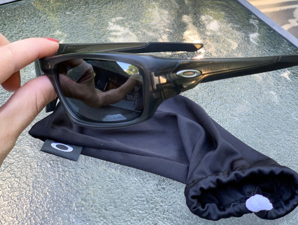 person holding up a black pair of oakley sunglasses with glass patio table and sunglasses storage bag in background