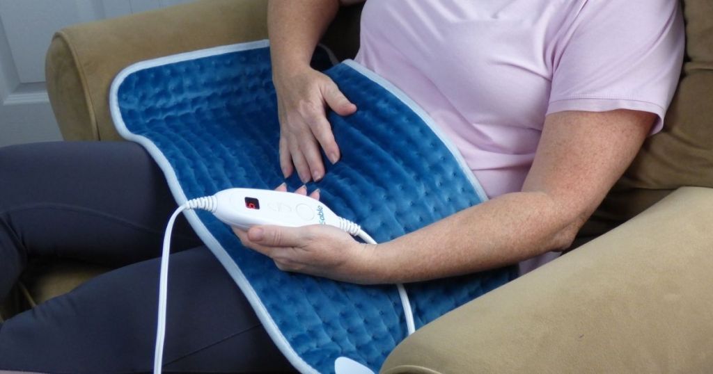 Woman sitting in arm chair, wearing a heating pad and holding the remote