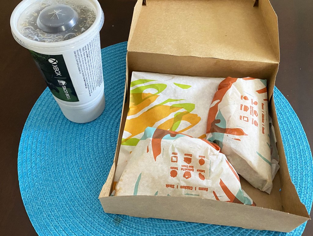 brown box with three taco bell items inside and drink cup next to it on blue placemat