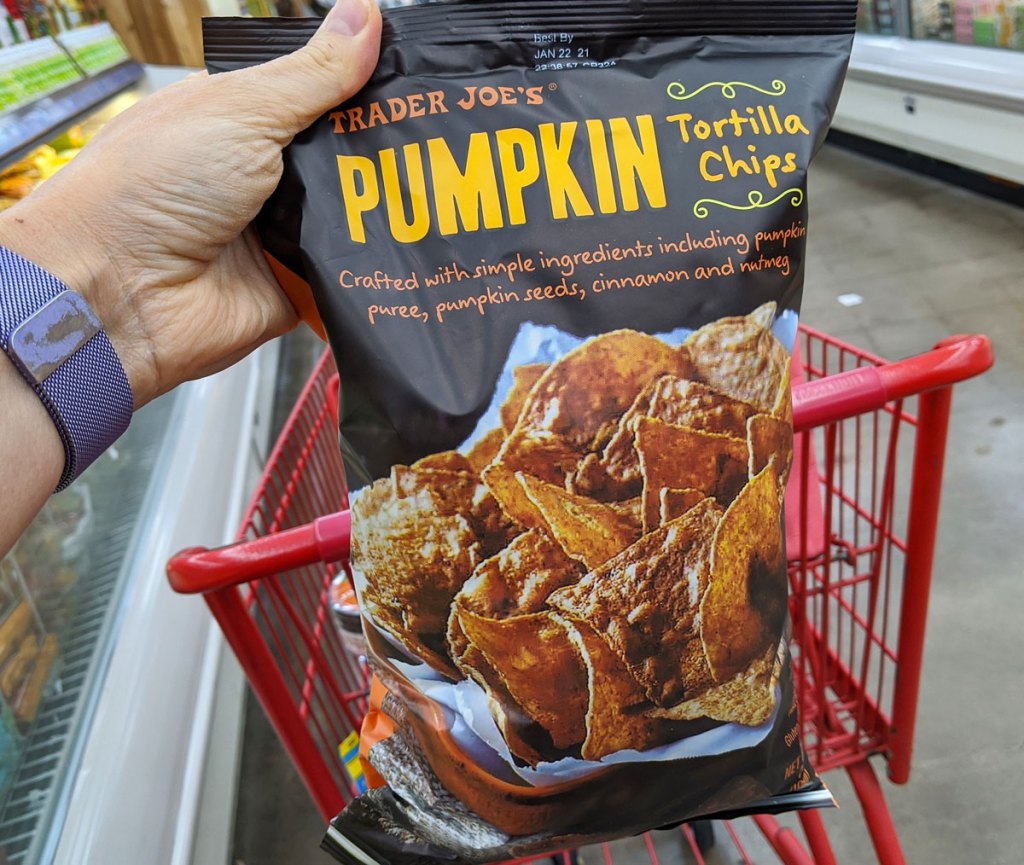 person holding up a black bag of pumpkin tortilla chips near red shopping cart