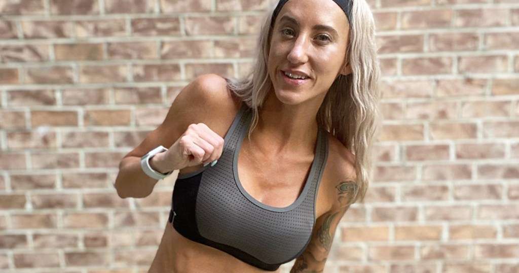 blonde haired woman standing in front of brick wall wearing a grey and black sports bra