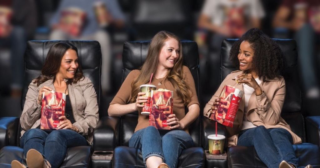 3 women sitting in movie theater holding drinks and popcorn talking and smiling