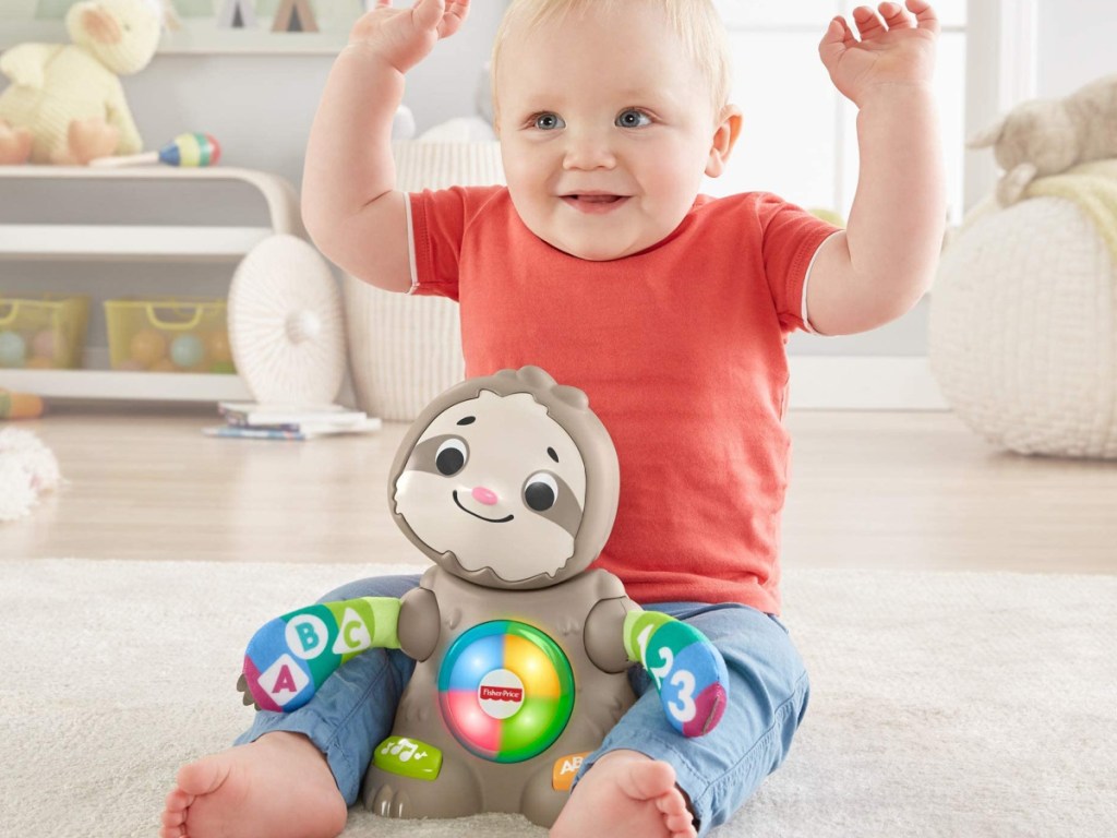 baby boy sitting on the floor with his hands up behind a fisher price sloth toy