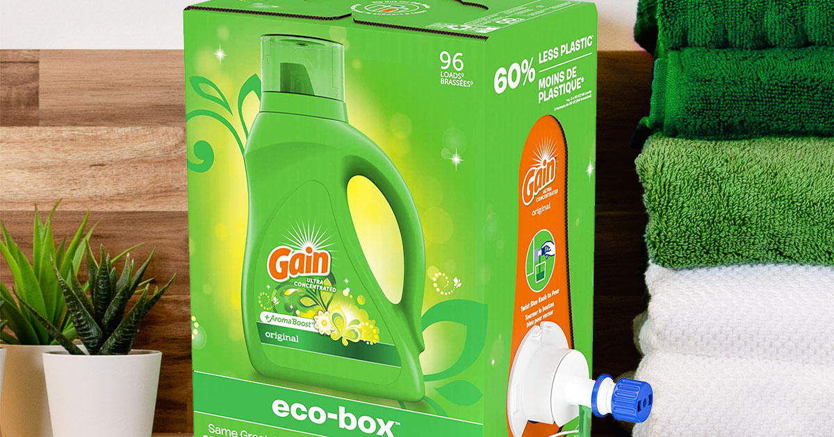 green box of gain laundry detergent near stack of folded towels and potted plants