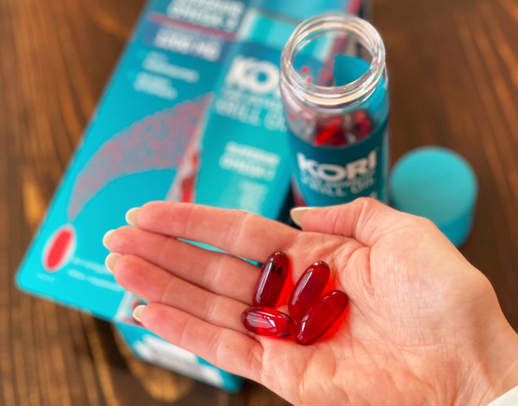 Kori Krill Oil bottle open on wooden table with hand holding red capsules