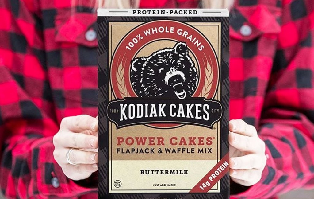 woman in red and black plaid shirt holding up box of kodiak cakes protein pancake mix
