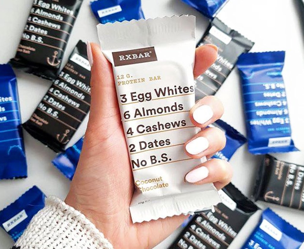 woman with white nails holding up a coconut chocolate RXBar protein bar with various other flavored bars behind it