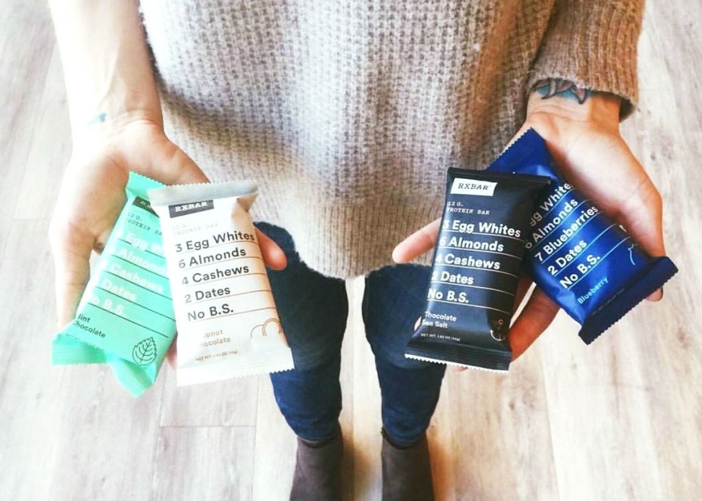 woman in cream sweater and jeans holding up two RXbar protein bars in each hand