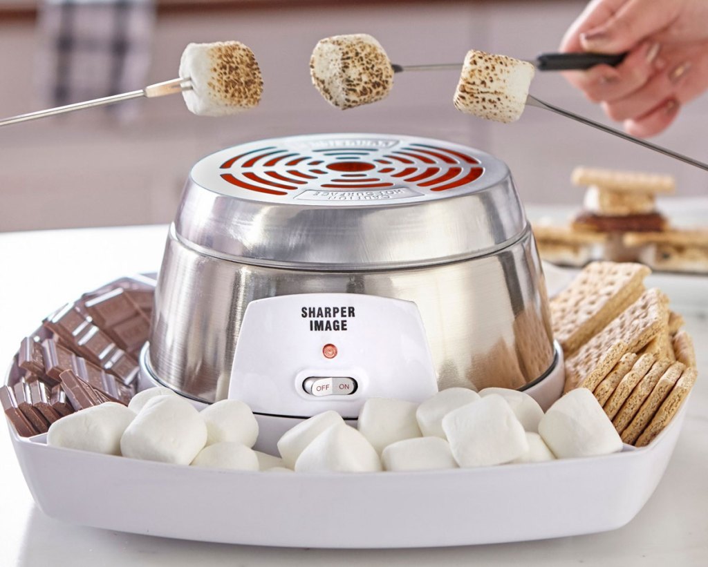 three people roasting marshmallows over a stainless steel smores maker with ingredients in tray around it