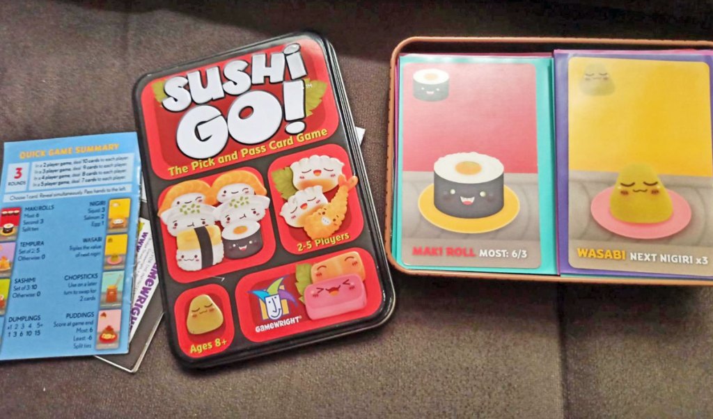 sushi go card game tin, instructions, and cards on a brown couch cushion