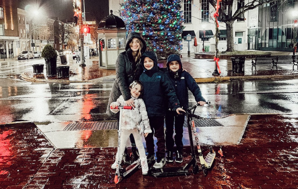 woman standing with kids on scooters downtown at night with rain and christmas lights