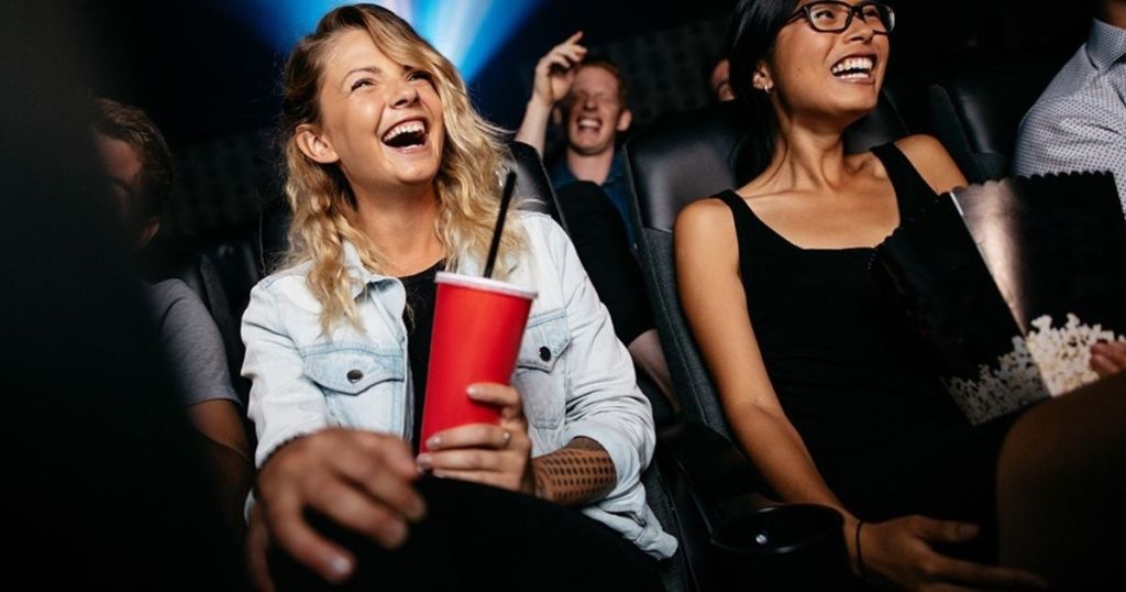 women sitting in movie theater holding drink and popcorn laughing