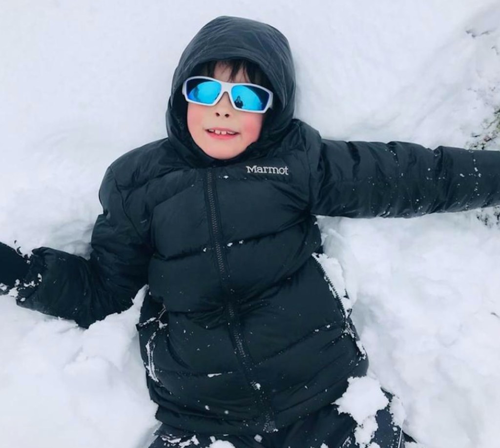 kid with navy winter jacket and blue lens sunglasses on laying in snow