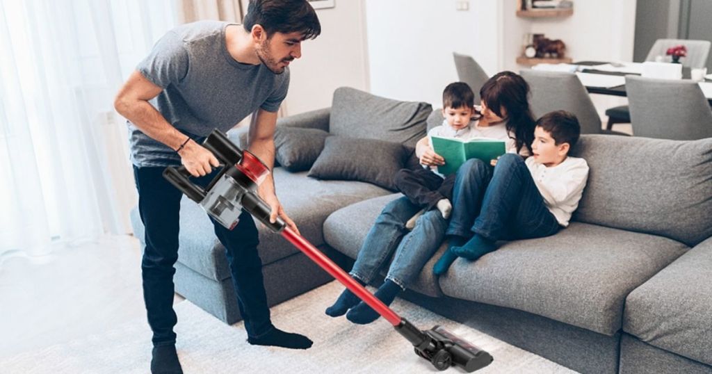 man using red and black stick vacuum to clean under couch with woman and 2 kids sitting on it
