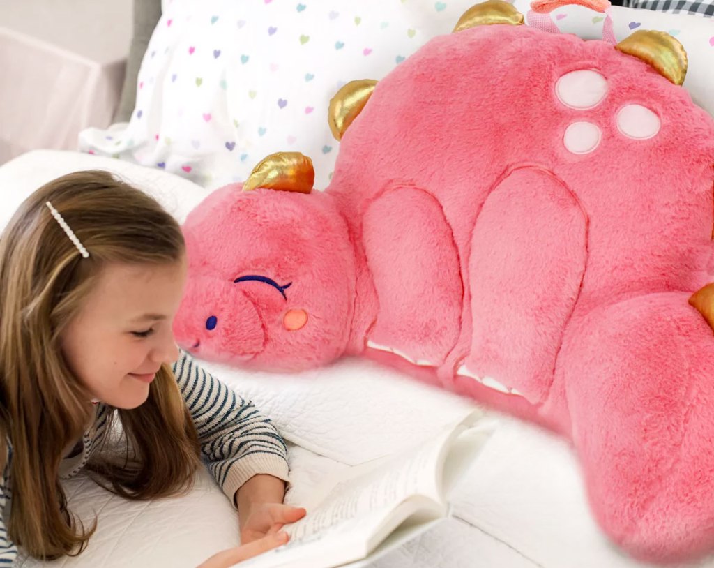 girl with brown hair laying on bed reading a book next to a pink dinosaur shaped backrest pillow