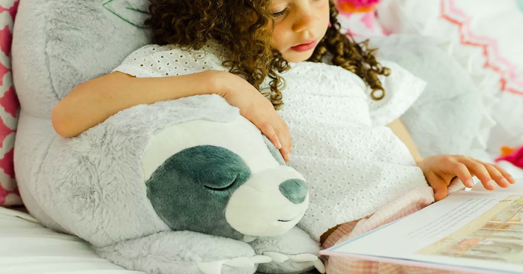 girl reading a book leaning against a grey sloth shaped backrest pillow