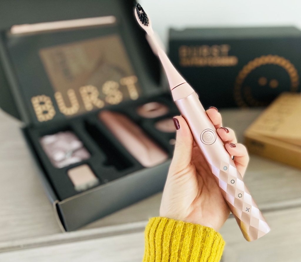 woman holding up a rose gold electric toothbrush with a starter kit in background