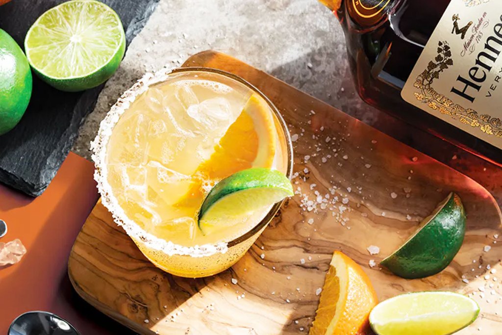 orange colored margarita with salt rim and lime on a wood cutting board with bottle of hennessy in background