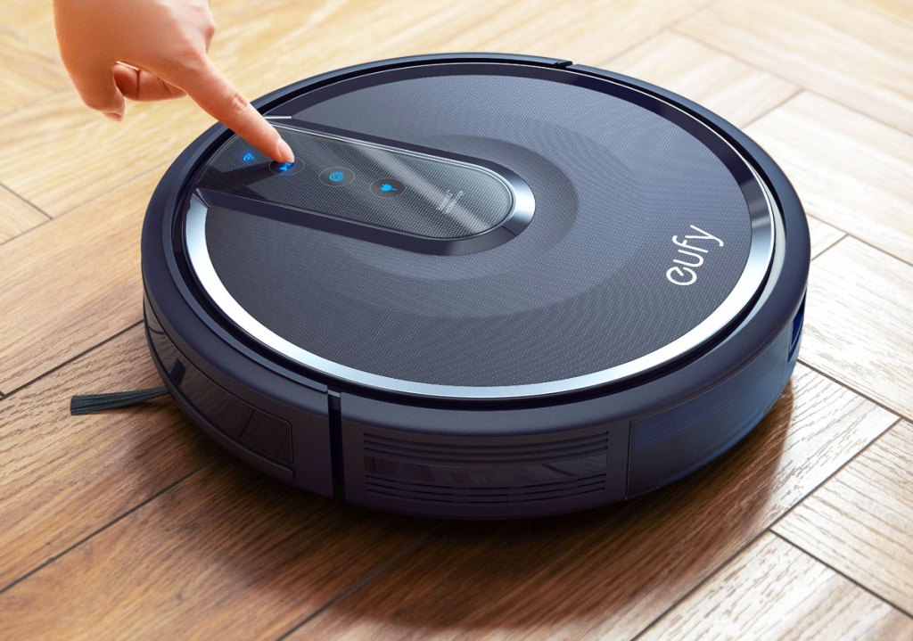 person pressing a button on a black eufy robotic vacuum on a wood floor