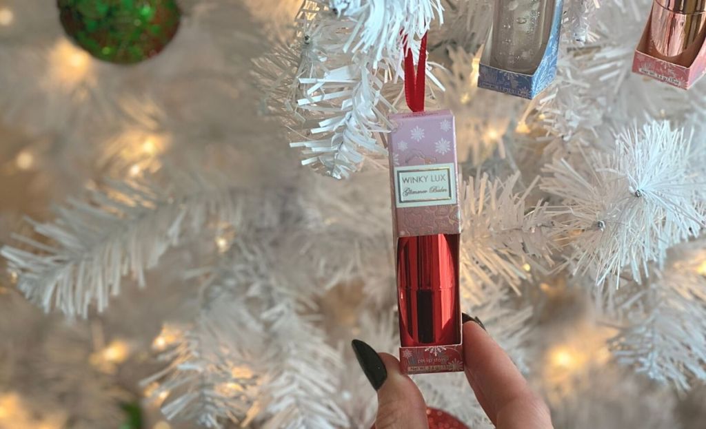A hand grabbing a winky lux holiday makeup ornament off of a white Christmas tree