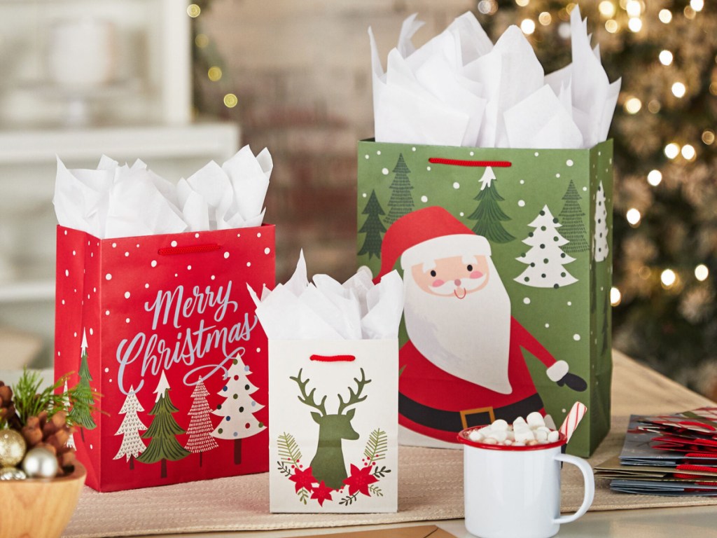 three gift bags with Christmas designs, cup of hot chocolate with marshmallows, and Christmas tree