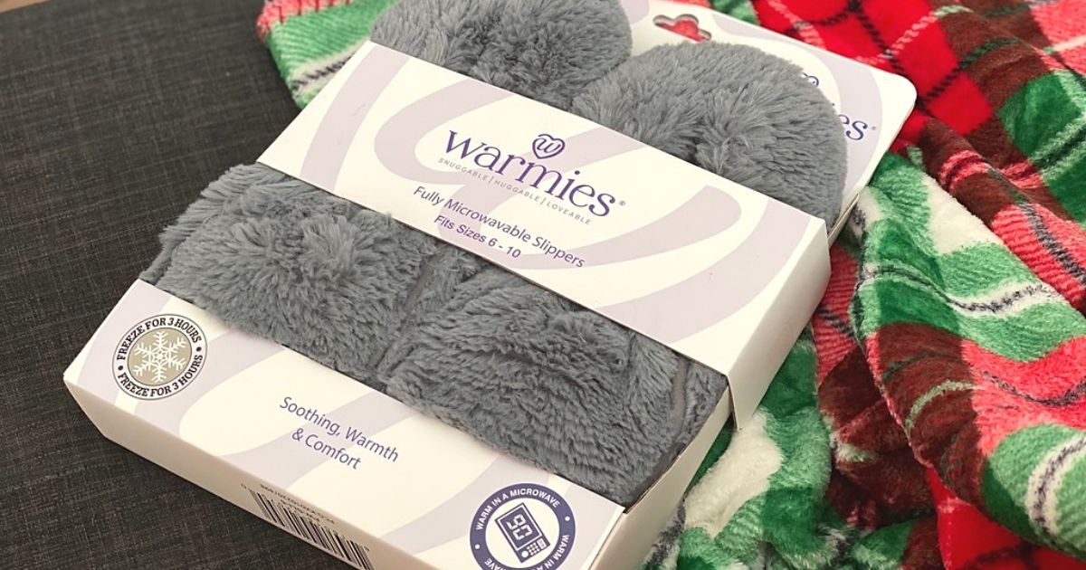 A pair of warmies slippers in box next to a plaid blanket