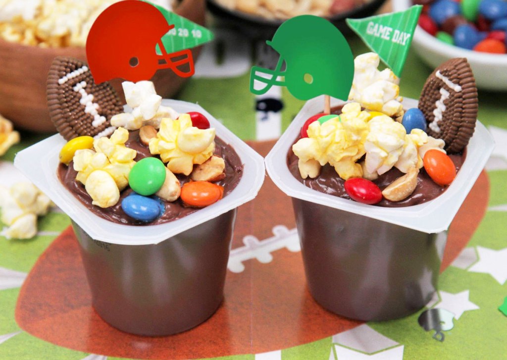 two chocolate snack pack pudding cups with popcorn, m&ms, and football helmet decorations on top