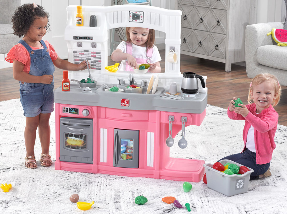 three girls in playroom playing with white and pink kitchen set with food items all over the floor