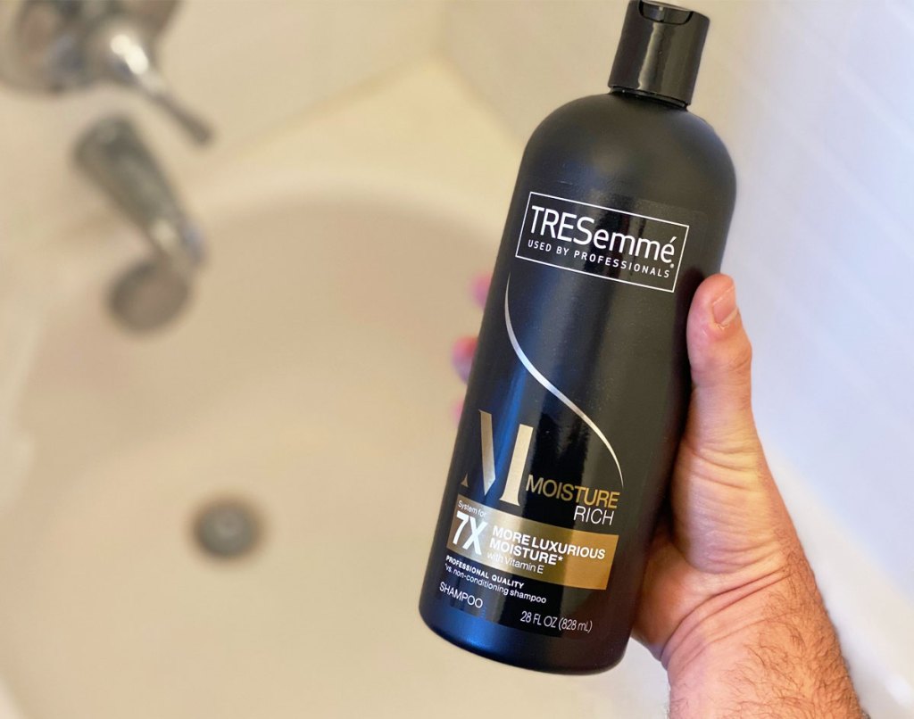 man holding up a black botle of tresemme shampoo with bathtub in background