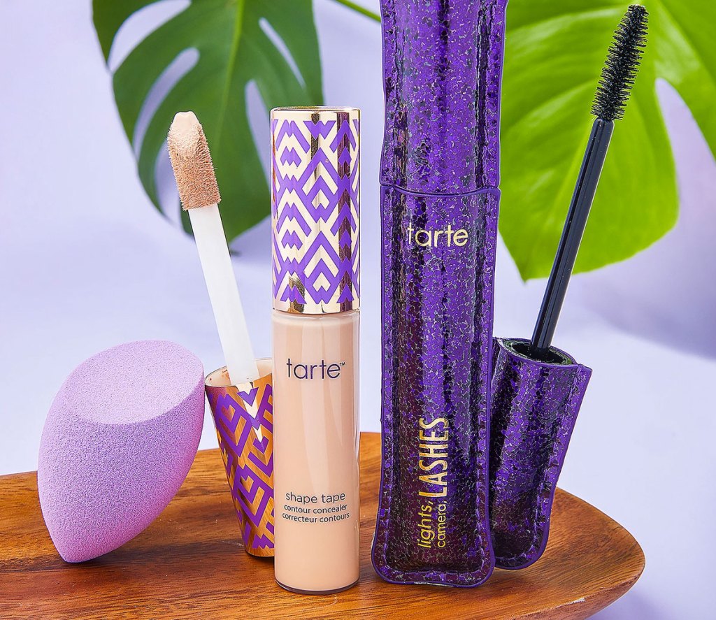 tarte shape tape, purple mascara tube, and purple blending sponge on a wood plate with tropical leaves in background