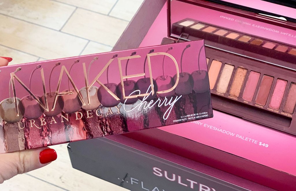 woman holding up an Urban Decay Naked Cherry eyeshadow palette next to an opened one on display