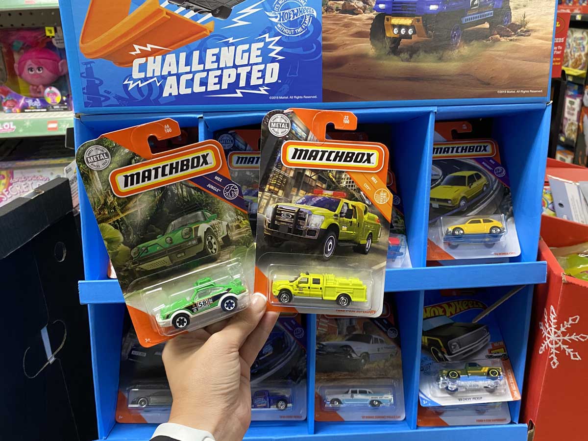 hand holding two matchbox cars in front of a store display of matchbox cars