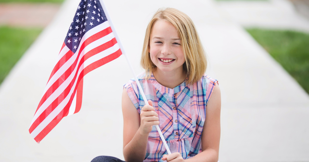 فتاة تجلس على الرصيف وابتسمت وتحمل علمًا أمريكيًا صغيرًا