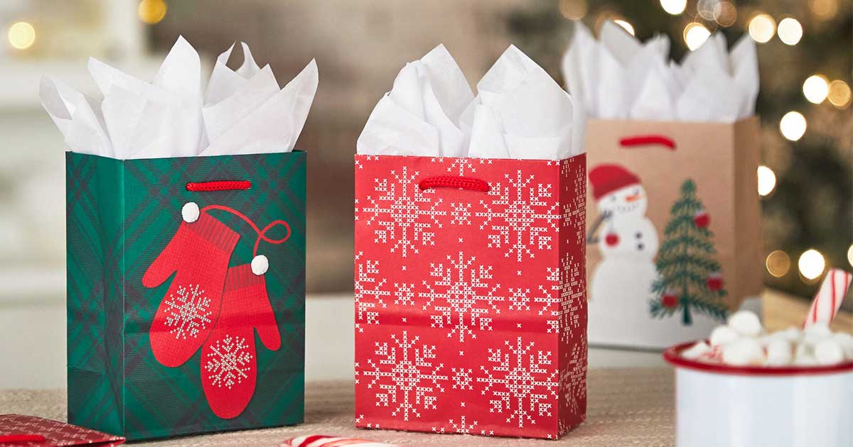 three Christmas themed gift bags next to hot cocoa on a table in front of a Christmas tree