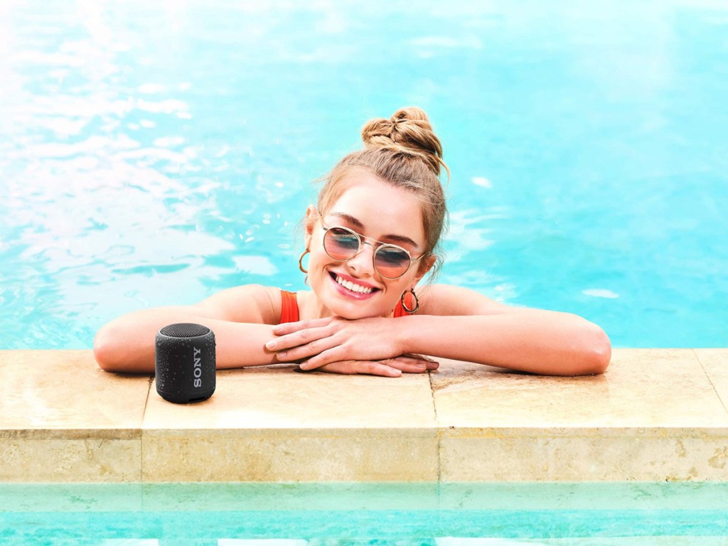 woman leaning on the side of a pool next to a sony mini speaker