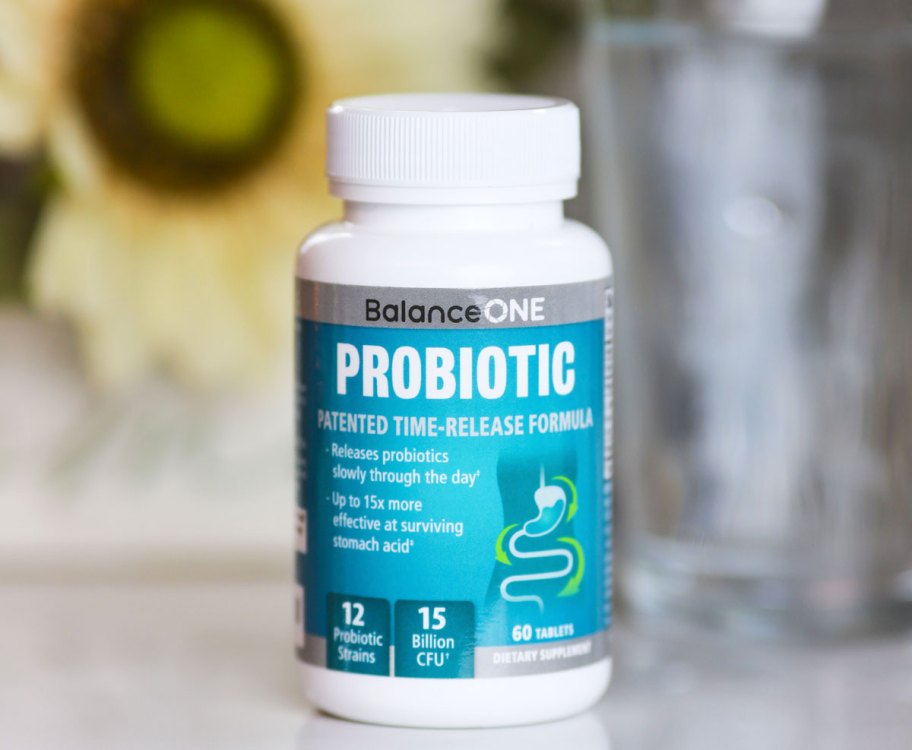 white and blue bottle of balance one probiotics sitting on a white table with flower and glass of water in background