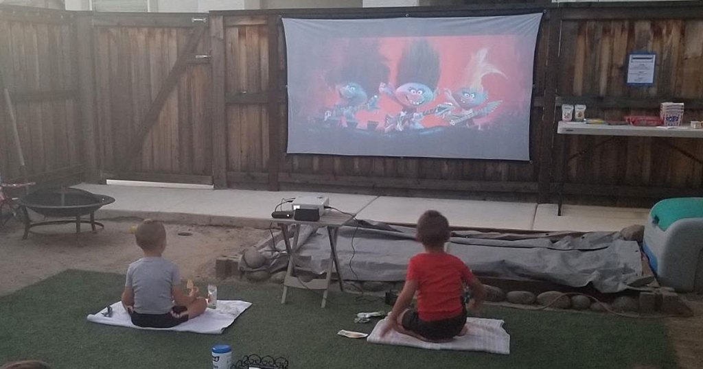 boys sitting in grass watching trolls movie projected on a screen on fence