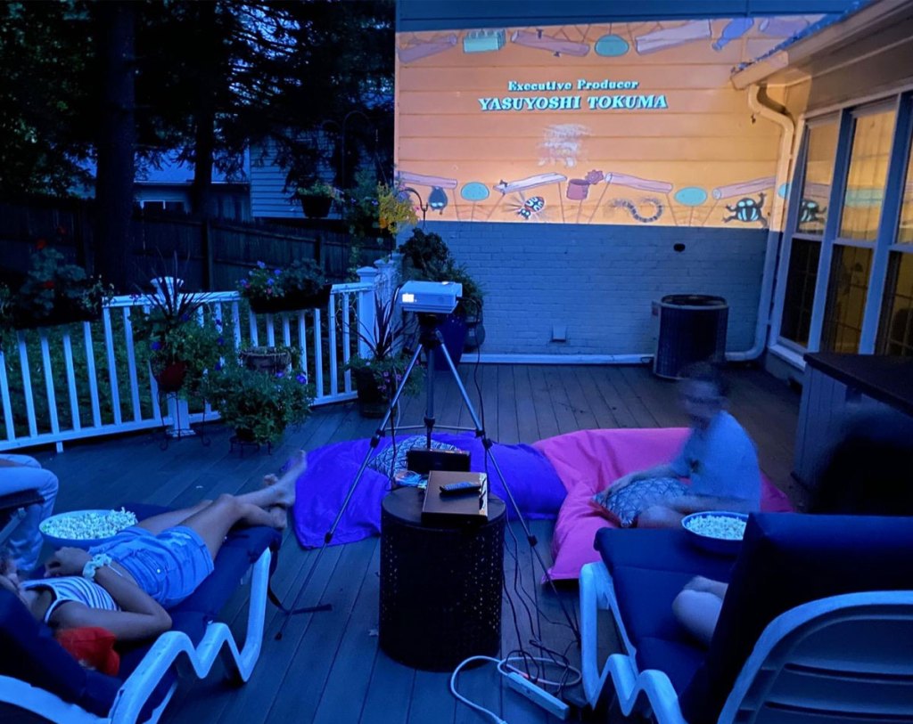 family laying on patio watching a movie projected on the side of the house with movie projector on tripod