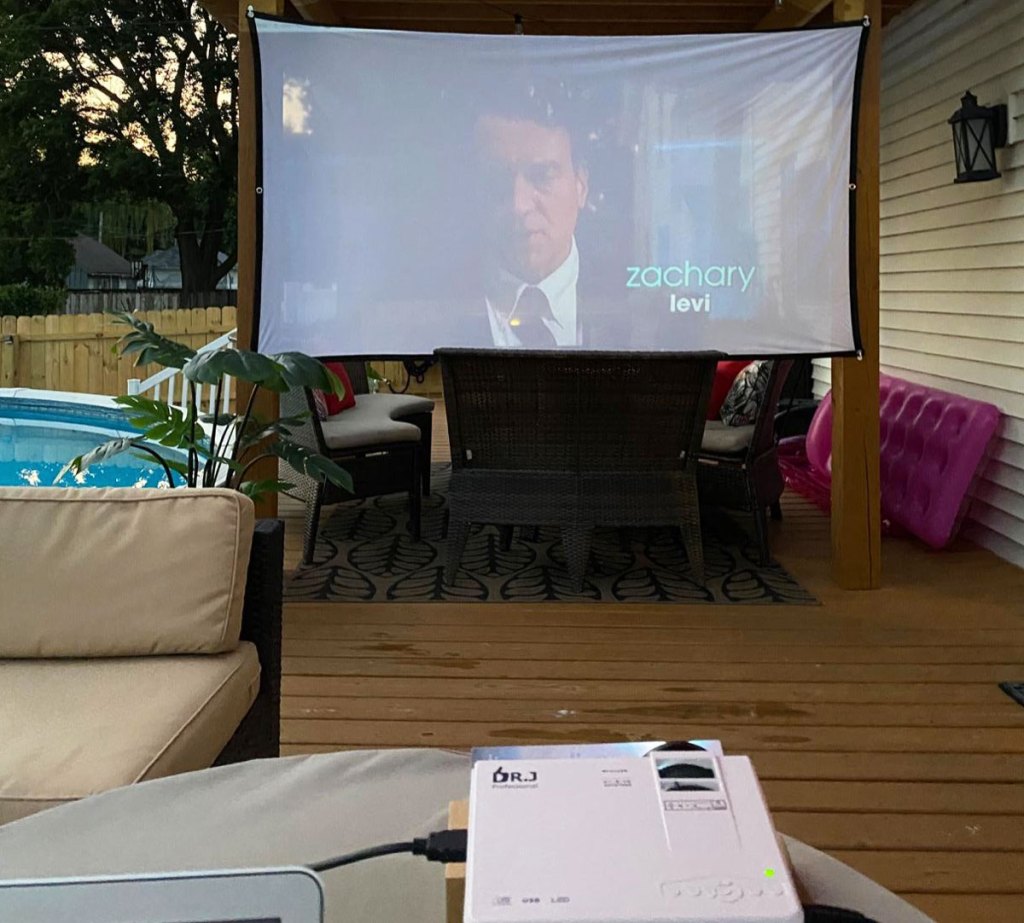 white movie projector projecting a movie on a large white screen attached to a patio outdoors