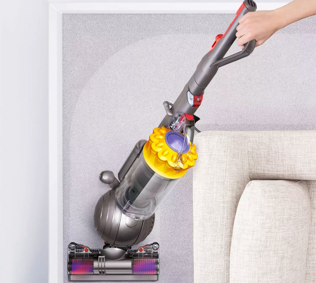 person pushing a grey and yellow dyson upright vacuum around the corner of a couch