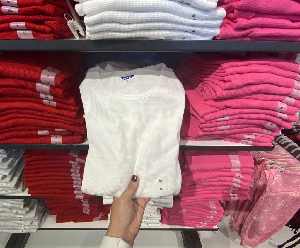 woman holding a white thermal tee from display shelves full of folded thermals in red, white, and pink colors