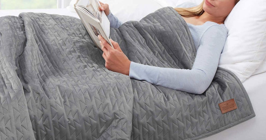 woman reading book in bed with a grey weighted blanket over her