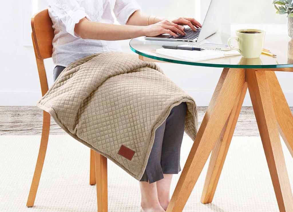 woman sitting at a table working from laptop with a tan weighted blanket on her lap