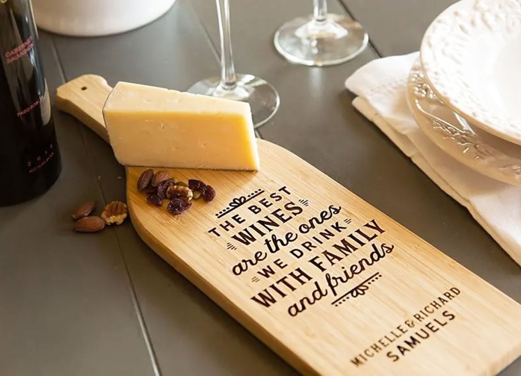 wine bottle shaped cutting board with personalized engraving on it near block of cheese and wine glasses