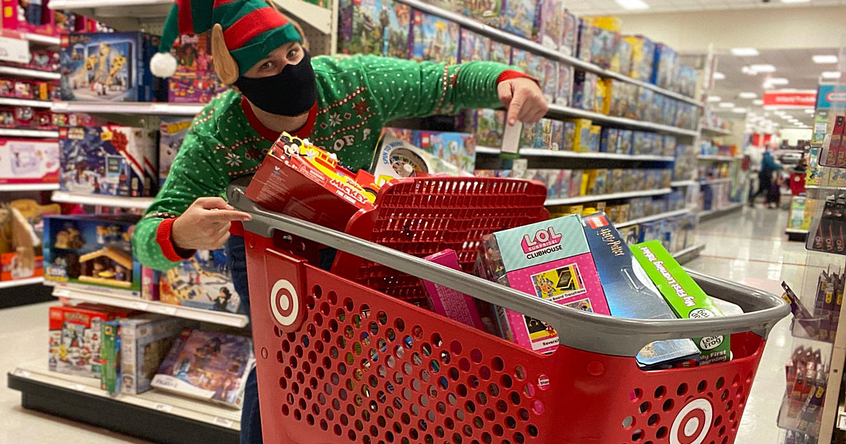 man in green sweater and elf hat with black face mask pointing at a red target shopping cart full of toys